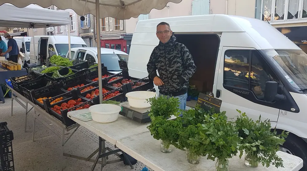 Marek derrière son étal de marché : cageots de tomates et de salades, bouquets de persil et balance sur la table.
